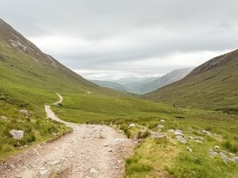 Grusvej slynger sig gennem grøn dal på West Highland Way, Skotland. Bølgende bakker på begge sider under overskyet himmel.