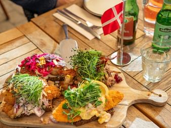 Träbräda med danska smörgåsar toppade med fisk, grönsaker och färska örter. En liten dansk flagga står i mitten.