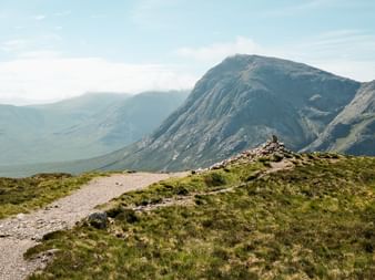 Grusvandrerute på West Highland Way med grønne græsskråninger til dramatiske bjergtinder under blå himmel i Skotland.