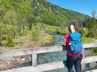 Two hikers with backpacks standing on a wooden bridge over a clear mountain stream in Austria, surrounded by green meadows and forested peaks.