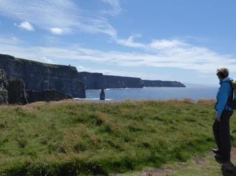 Vandrer i blå jakke med rygsæk står på græsklædt klippe med udsigt over dramatiske havklipper og hav under skyet himmel.