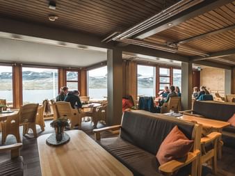 Interiør i DNT Finsehytta hytte med tremøbler, mørke sofaer med oransje puter og store vinduer med utsikt over innsjø og fjell.