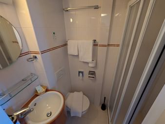 Kompakt badeværelse på MS Rigoletto med hvide fliser, glasbruser, toilet, vask med spejl og håndklæder hængende på væggen.