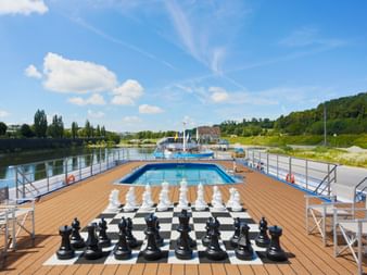 Soldæk på MS SE-MANON med kæmpeskakspil på skakternsgulv, swimmingpool, trædæk og flodudigt med grønne bakker.