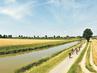 Gruppe af cyklister på en asfalteret sti langs en kanal i Po-regionen. Gyldne hvedefelter strækker sig over det flade landskab.