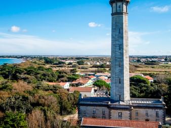 Højt stenfyrtårn med udsigt over La Rochelles kystlinje med røde teglstenstage, grøn vegetation og blåt ocean i baggrunden.