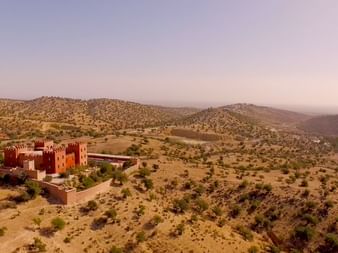 Atlas Kasbah Ecolodge i marokkanske bjerge Luftfoto af Atlas Kasbah Ecolodge, en traditionel rød fæstningslignende bygning omgivet af tørre bakker i Marokko.