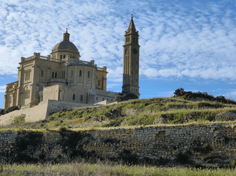 Ta' Pinu basilika på Gozo med cremefarvede stenfacade, stor kuppel og højt klokketårn, beliggende på en bakke med stenmure og vegetation.