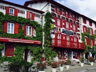 Row of traditional white Basque buildings with distinctive red shutters and ivy-covered walls. Hotel-restaurant signage visible on ground floor.