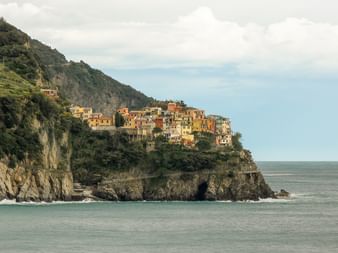 Colorful houses of a Cinque Terre village perched on a steep rocky cliff overlooking the Mediterranean Sea under a cloudy sky.