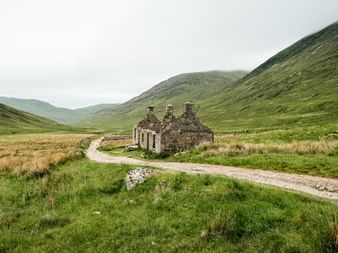 Stenruiner af et gammelt hus ved en grusvej i en grøn skotsk dal. Bakkede åse rejser sig på begge sider under en overskyet himmel.