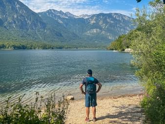 Vandrare med ryggsäck på stenstrand vid alpin sjö i Juliska Alperna, Slovenien. Berg och skog synliga i bakgrunden.