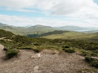 Grusvej gennem skotske højlande på West Highland Way med grønne bakker, bjerge og fjern sø under skyet himmel.