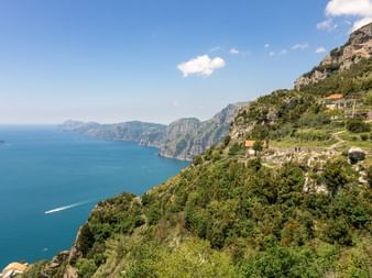 Panoramaudsigt over Amalfikysten med dramatiske klipper dækket af grøn vegetation, traditionelle huse på terrasserede skråninger og turkisblåt hav.