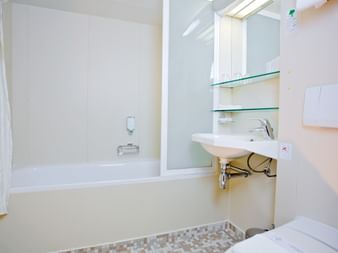 Moderne hvidt badeværelse på MS SE-MANON med badekar, vægmonteret vask, glashylder, spejl og mosaikbort.