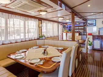 Interiør af MS Amore krydstogtskibs spisestue med dækket træbord, beige siddepladser, store vinduer med gardiner og køkkenområde.