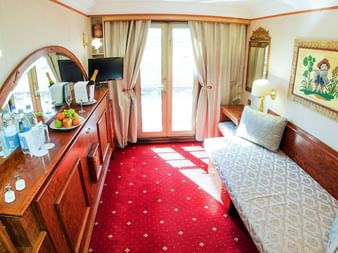 Kompakt skibskahyt med enkeltseng, trækommode med spejl, frugtkurv og balkondør med gardiner. Rødt mønstret gulvtæppe dækker gulvet.