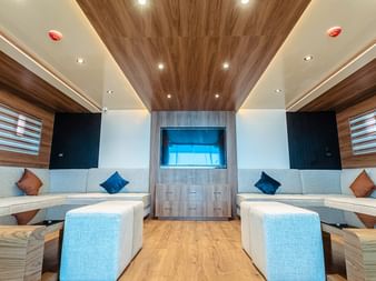 Moderne bådinteriør med hvide polstrede sæder langs begge vægge, farverige puder, træpaneler og blå accentbelysning.