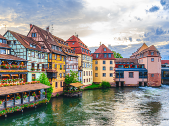 Traditionelle bindingsværkshuse i livlige farver langs en kanal i Strasbourg. Bygninger med blomstersmykkede terrasser afspejles i vandet.