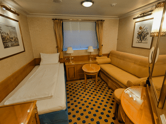 Kompakt kahyt på MS Casanova med enkelseng, beige sofa, rundt bord, vindue med gardiner og blåt mønstret tæppe.