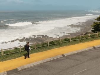 Pilgrim with backpack walking on yellow-marked coastal path along the Portuguese Camino. Rocky coastline with waves and wooden fence visible.