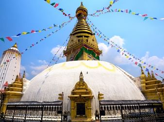 Swayambhunath Stupa med bedeflag Swayambhunath Stupa i Nepal med hvid kuppel, gylden spir og farverige bedeflag der stråler fra toppen mod blå himmel med skyer.