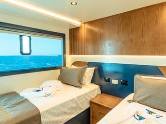 Moderne to-sengskahyt ombord på High Wave med træpaneler, blå accenter og stort vindue med havudsigt.