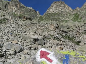 Rocky mountain terrain with colorful trail markers painted on stone. Red and white directional arrow with blue and yellow paint marks visible on boulder.