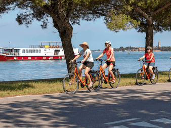 Fire cyklister på orange cykler på en strandvej. En rød og hvid krydstogtbåd ses på det blå vand, træer indramme scenen.