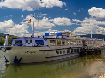 Hvidt flodkrydstogtskib ved navn Olympia fortøjet ved kaj på floden Mosel, med bakker og blå himmel med hvide skyer i baggrunden.