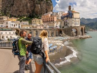 Tre vandrere med rygsække står på et udsigtspunkt og ser ud over kystbyen Atrani på Amalfikysten, med strand og Middelhavet.