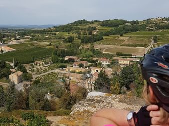 Cyclist with black helmet viewing panoramic landscape of Côtes du Ventoux wine region with vineyards, village houses, and rolling hills.