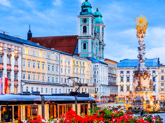 Historisk plads i Linz med barokbygninger, blåt kirketårn, udsmykket Treenigheds søjle og røde blomster i forgrunden.