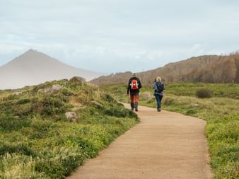 Two hikers with backpacks walking on a sandy path through green countryside on the Portuguese Camino, with a mountain visible in the distance.