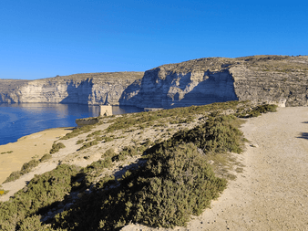 Grusvej langs Middelhavets kyst på Gozo med dramatiske kalkstensklipper, sandstrand og blåt hav under klar himmel.
