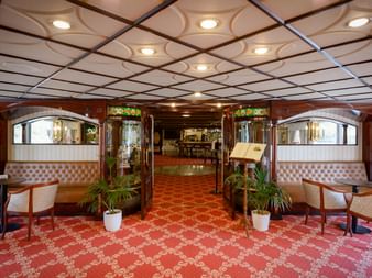 Elegant salon interiør på MS Prinzessin Katharina med rødt mønstret tæppe, hvide læderstole, dekorativt loft og planter.