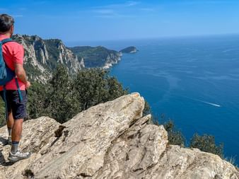 Hiker with backpack standing on rocky cliff overlooking the blue Mediterranean Sea and Ligurian coastline with dramatic cliffs and islands.