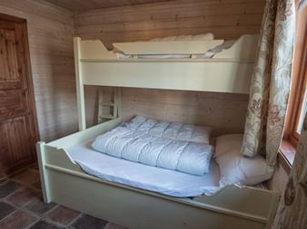 Hvid køjeseng med hvidt sengetøj i et træhytterum. Naturlige trævægge og loft, trædør, vindue med gardiner og flisegulv.
