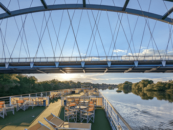 Udsigt fra MS Casanovas soldæk under passage af en moderne skråstagsbro. Liggestole og borde synlige på dækket med flod og træer forude.