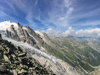 Panoramic view of Mont Blanc's snow-covered peaks and glacier with rocky foreground and green alpine valley below under blue sky with clouds.
