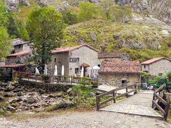 Stenhuse med terracotta-tage i en bjerglandsby i Picos de Europa. En træbro krydser en bæk med klippefyldte skråninger i baggrunden.