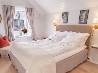 Lyst hotelværelse med beige dobbeltseng, hvidt sengetøj, væglamper, vindue med gardiner, lænestol og trægulv på Husum Hotel.