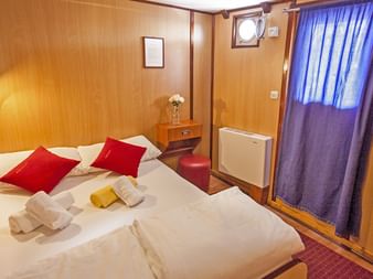 Hyggelig skibskahyt på MS Azimut med dobbeltseng, røde puder, træpaneler, blå gardiner, koøje og rød stol.