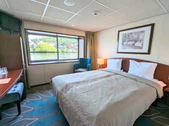 Moderne skibskahyt på MS SE-MANON med dobbeltseng, stort vindue mod flod, skrivebord, lænestol og indrammet kunst på væg.