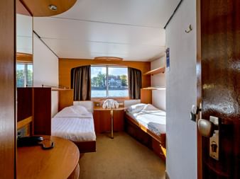Kompakt skibskahyt på MS Olympia med to enkeltsenge, træpaneler, rundt vindue med gardiner og indbyggede møbler.