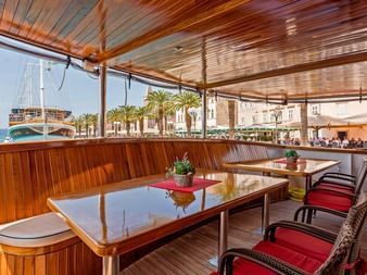 Overdækket spisedæk på MS Amore med træborde, røde stole og panoramavinduer med udsigt over kroatisk havn med palmer.