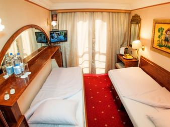 Kompakt tomandskabysse med to enkeltsenge, træmøbler, ovalt spejl, rødt tæppe, hvide gardiner og væg-TV på Donau-skib.