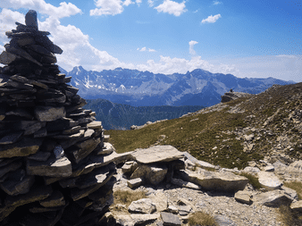 Stenvarde på klippefyldt alpint terræn med snedækkede bjergtinder i det fjerne under blå himmel med hvide skyer på Tour du Queyras.