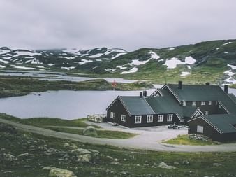 Mørk træhytte Geiterygghytta med grønt tag ved en bjergsø i Hardangervidda. Snepletter på grønne bakker under overskyet himmel.