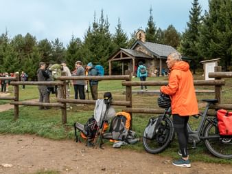 Syklist i oransje jakke med sykkel og ryggsekker ved tregjerdet, mens gruppe pilgrimer samles ved rustikt herberge.